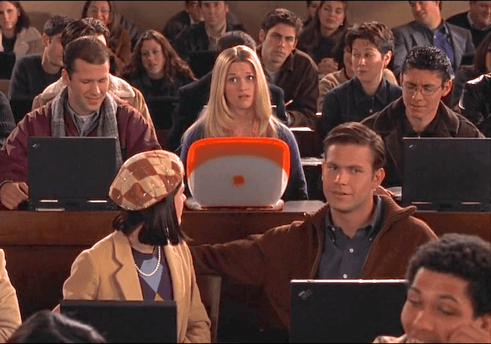 A screenshot of the movie "Legally Blonde", pictured is Elle Wood in a lecture hall using a Mac as she is surrounded by other individuals with simple black laptops.